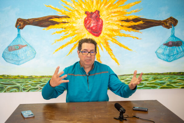 A photo of Abel Oriz during an interview. He is sitting at a brown wooden table with a mural behind him. The mural includes a human heart in a starburst.