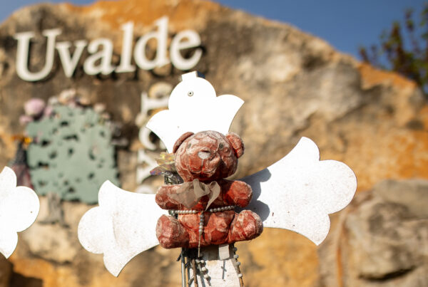 A faded teddy bear is tied to a small white cross. In the background is the broken city limits sign of Uvalde, Texas. The Texas is tilted perpendicular.