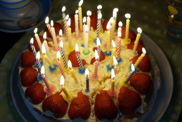 A photo of a homemade birthday cake topped with white icing, lots of strawberries and many lit candles.