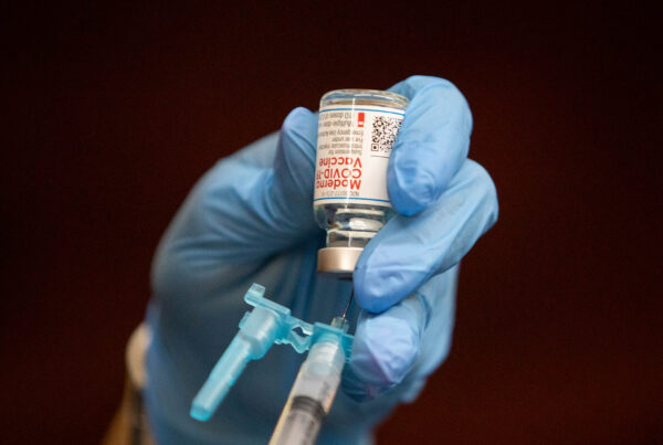 A close-up of a hand wearing a blue medical glove drawing a vaccine from its bottle using syringe