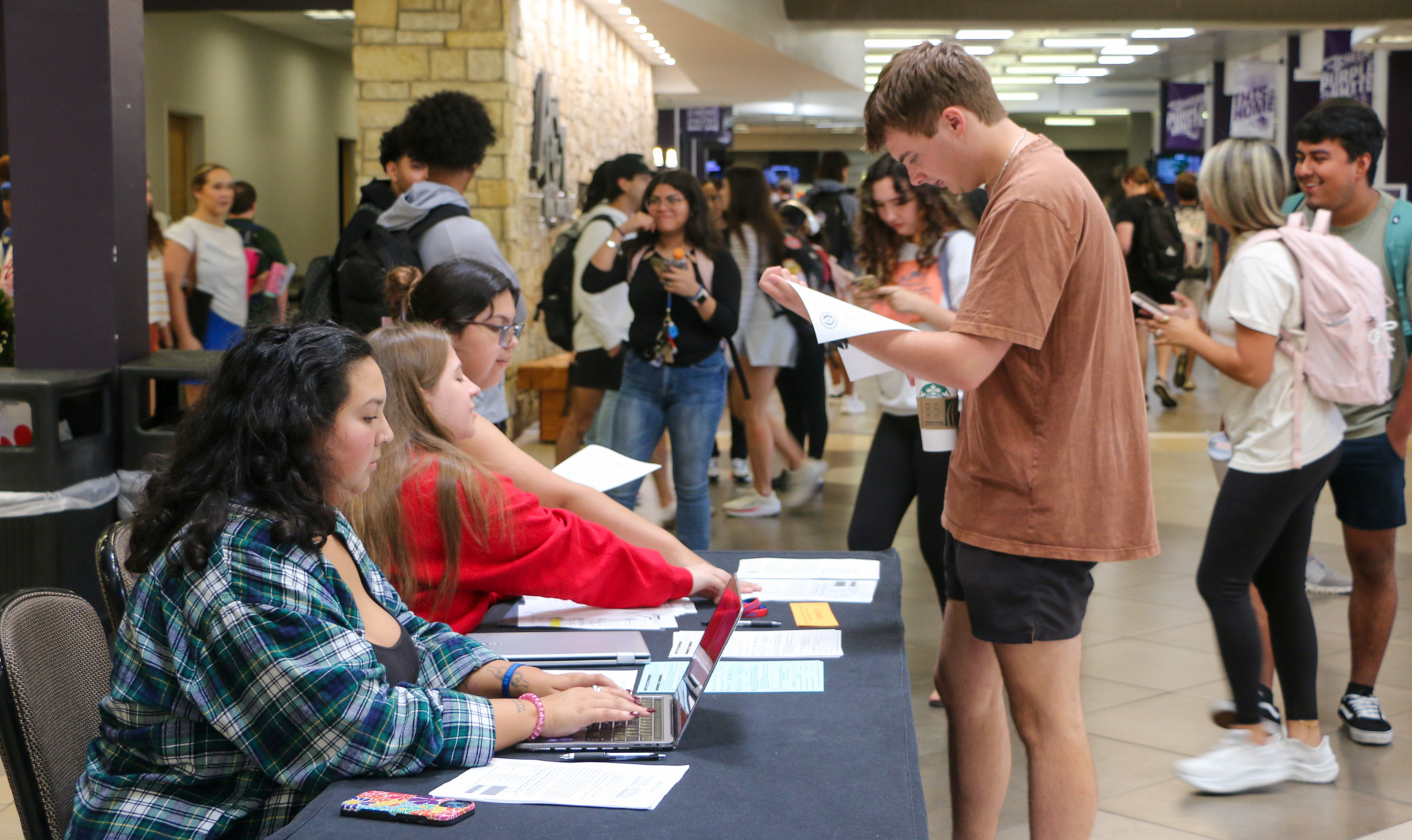 A student looks over a sheet of paper in front of a table where three other students sit with other information and laptops in a busy hallway.