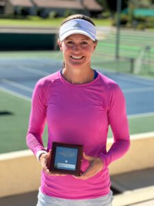 A woman in a long-sleeved pink shirt stands in front of a tennis court with an award.