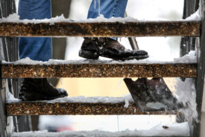 A closeup of boots walking down outdoor steps that have a layer of show and ice on them, with a shovel scraping one step