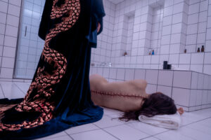 A still from the movie shows a person in a dark, ornate robe standing over another person with crude stitches down their spine lying on the floor of a white tiled bathroom.